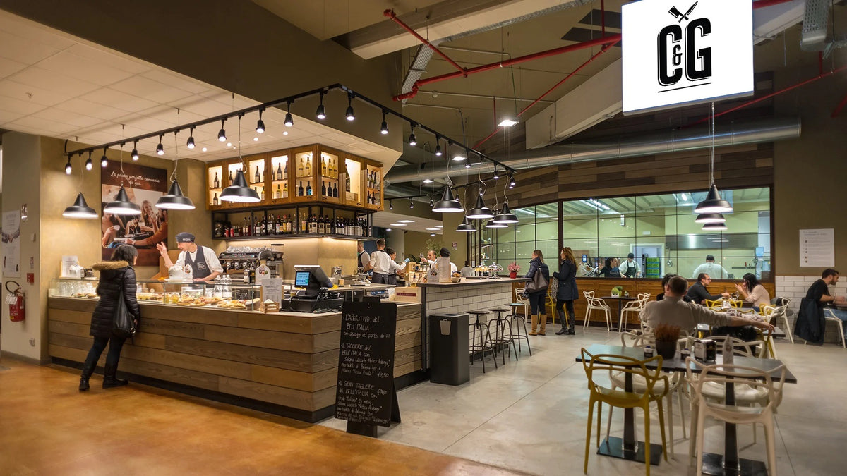 A bustling indoor cafe and bakery scene with a long wooden counter, hanging pendant lights, customers seated at tables, and a large white hanging sign displaying the 'CG' logo.